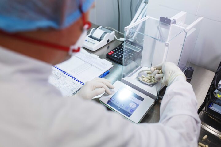 over-the-shoulder picture of a man in a lab coat and protective gear running tests on pharmaceutical products in a testing box.