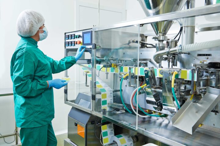 pharmaceutical equipment with equipment operator in full body scrubs, gloves, hairnet, and mask operating machine