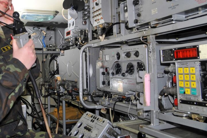 a wall of Military technology equipment with a soldier in the left of the frame listening to the hearing piece while holding a piece of equipment