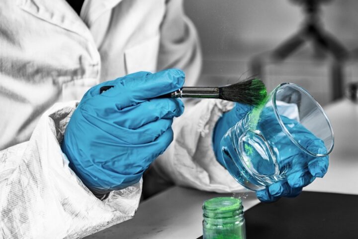 Close up of person in a white lab coat with protective gloves dusting a glass with testing substance.