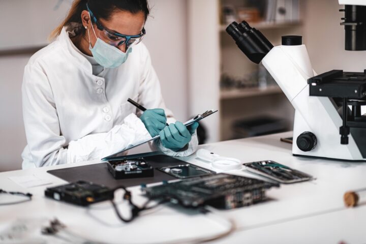 A female scientist with a white lab coat, face mask, protective glasses, and gloves taking notes with a microscope in frame.